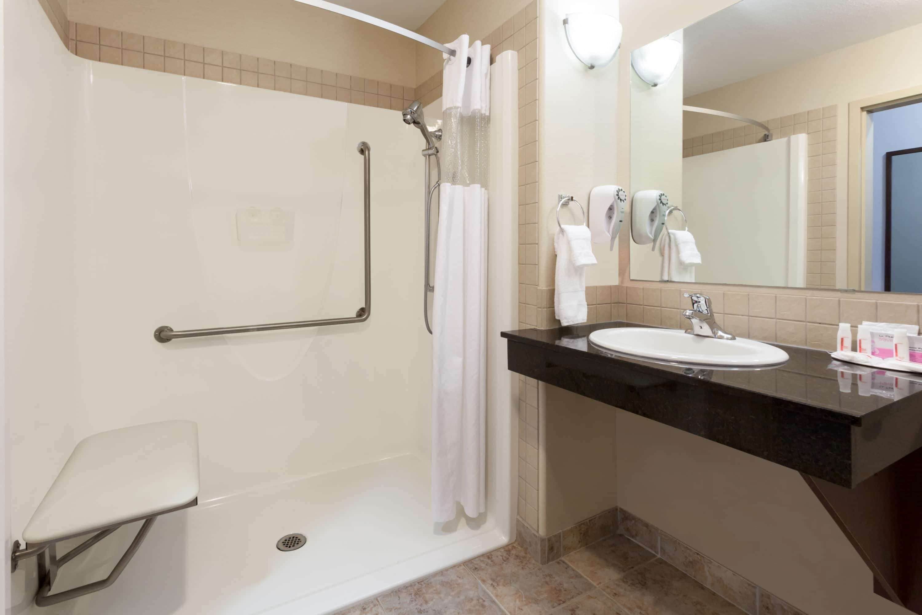 Un baño con ducha a nivel, barras de apoyo, lavamanos y espejo.