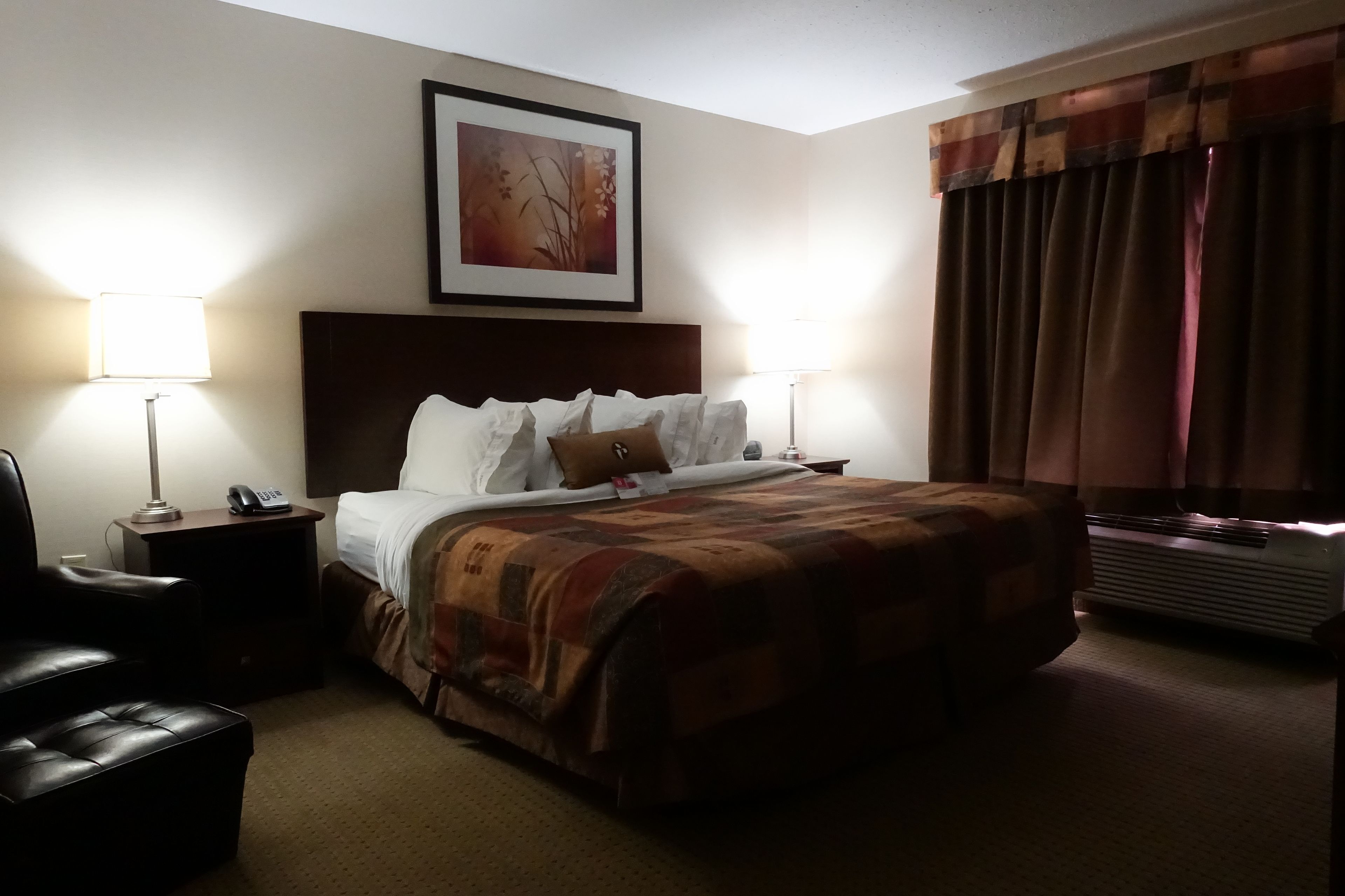 Habitación de hotel con cama, dos lámparas de noche, una silla, un cuadro en la pared y una ventana con cortinas.
