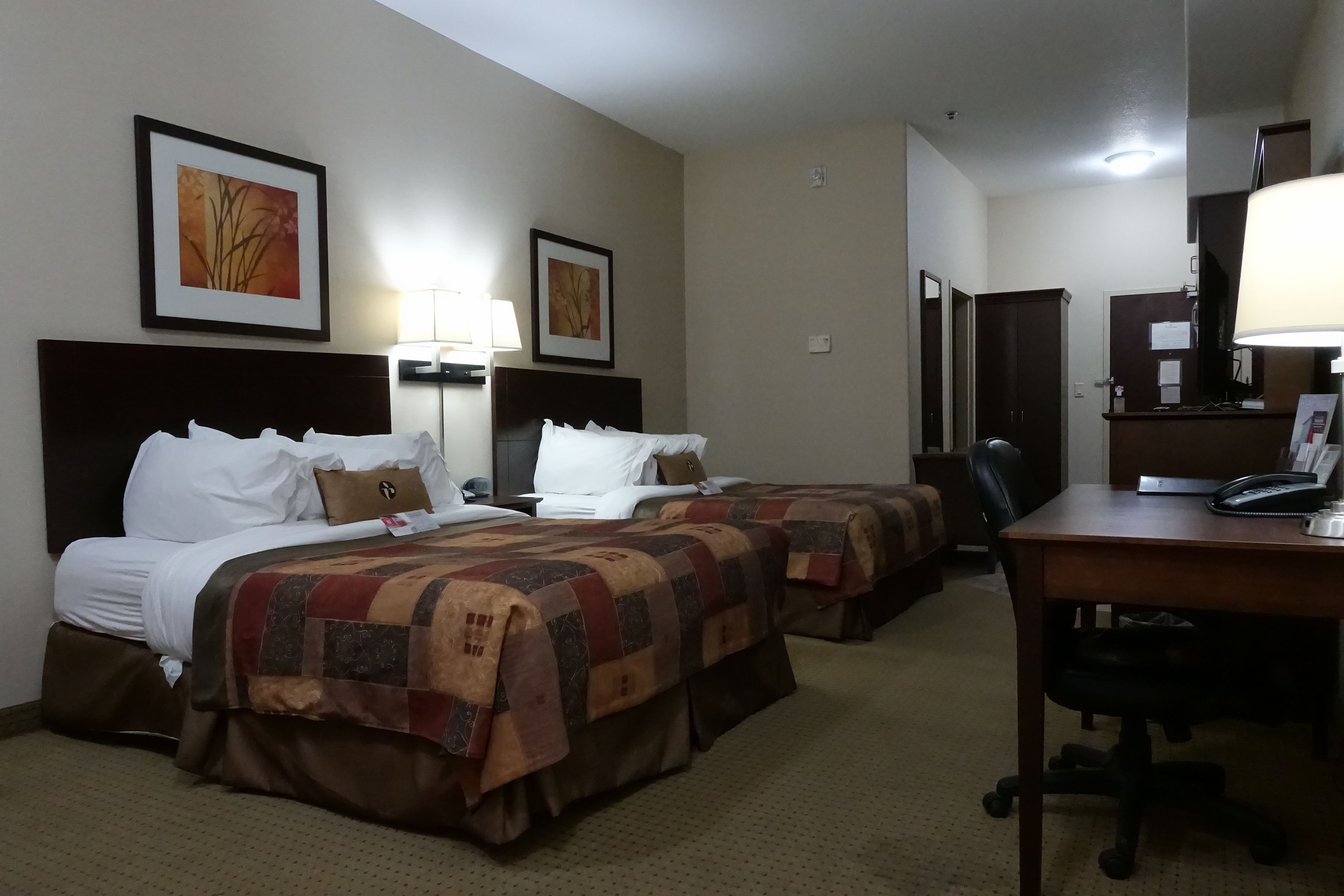 A hotel room with two beds, a desk, and a chair. There are framed pictures on the wall, a lamp, and a telephone on the desk.