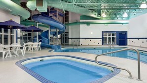 Una piscina interior con suelo de baldosas azules y blancas, un tobogán azul y sillas blancas alrededor de la zona de la piscina.