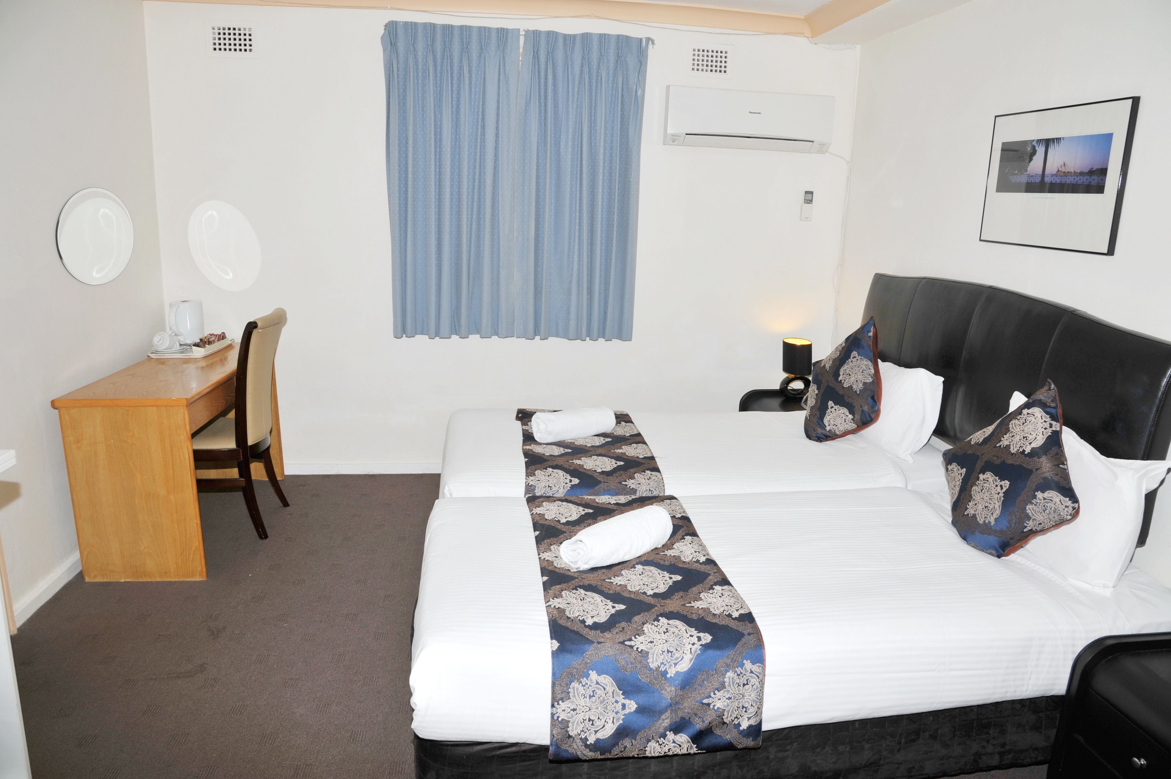 Quarto de hotel com cama, escrivaninha, cadeira, janela com cortinas, ar condicionado e quadro na parede.