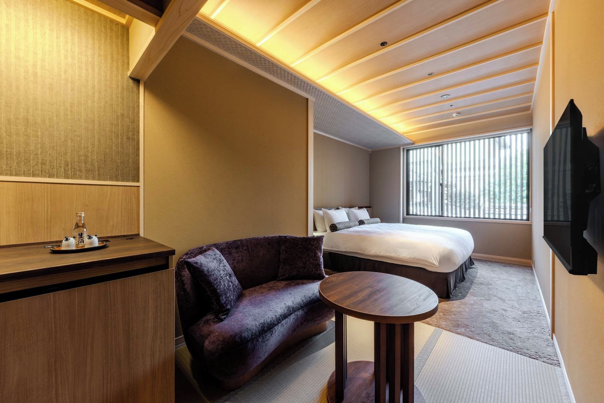 A hotel room with a bed, a seating area with a chair, a small round table, a wooden desk, and a TV mounted on the wall.