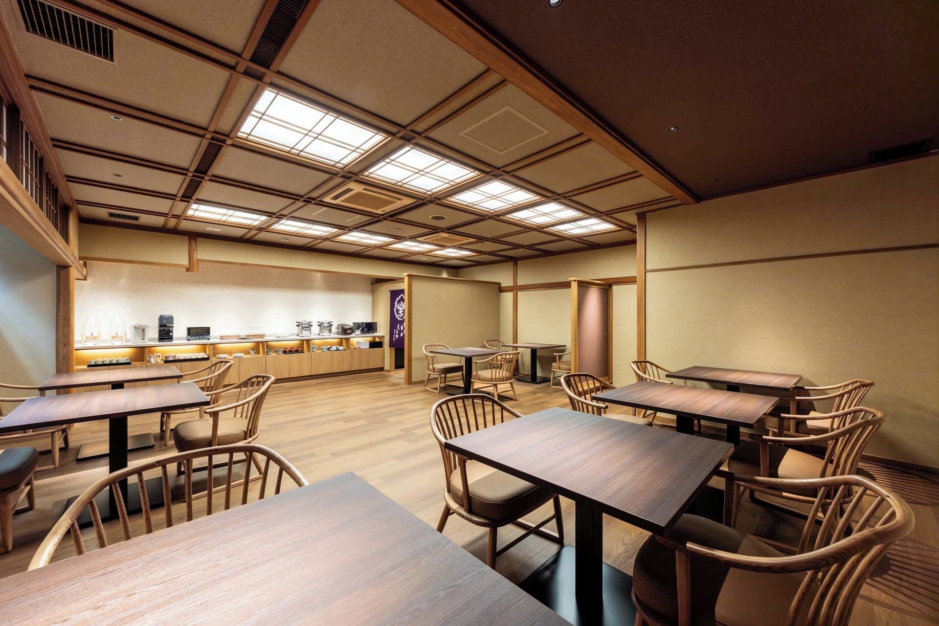 A spacious dining area with wooden tables and chairs, a kitchen in the background, and a high ceiling with skylights.