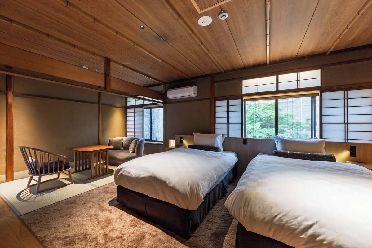 A hotel room with two beds, a chair, a small table, and large windows.