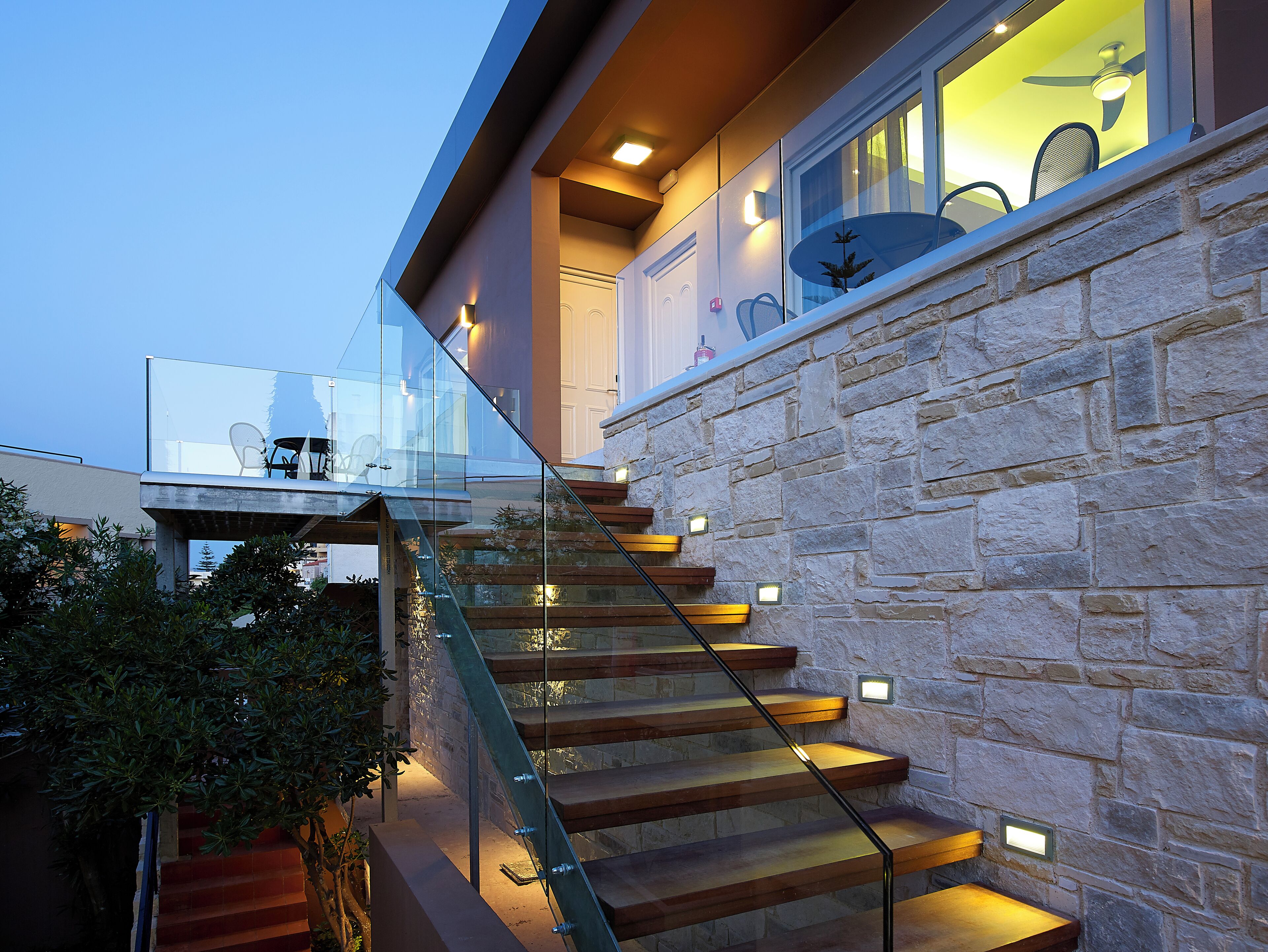 Una escalera moderna con barandillas de cristal y peldaños de madera, que conduce a un balcón con mobiliario exterior.