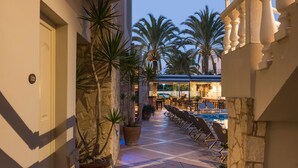 A corridor with a pool area, palm trees, and a bar in the background. - Elotis Suites (Chania)
