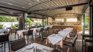 An outdoor dining area with tables and chairs, a stone wall, and a kitchen in the background. - Elotis Suites (Chania)