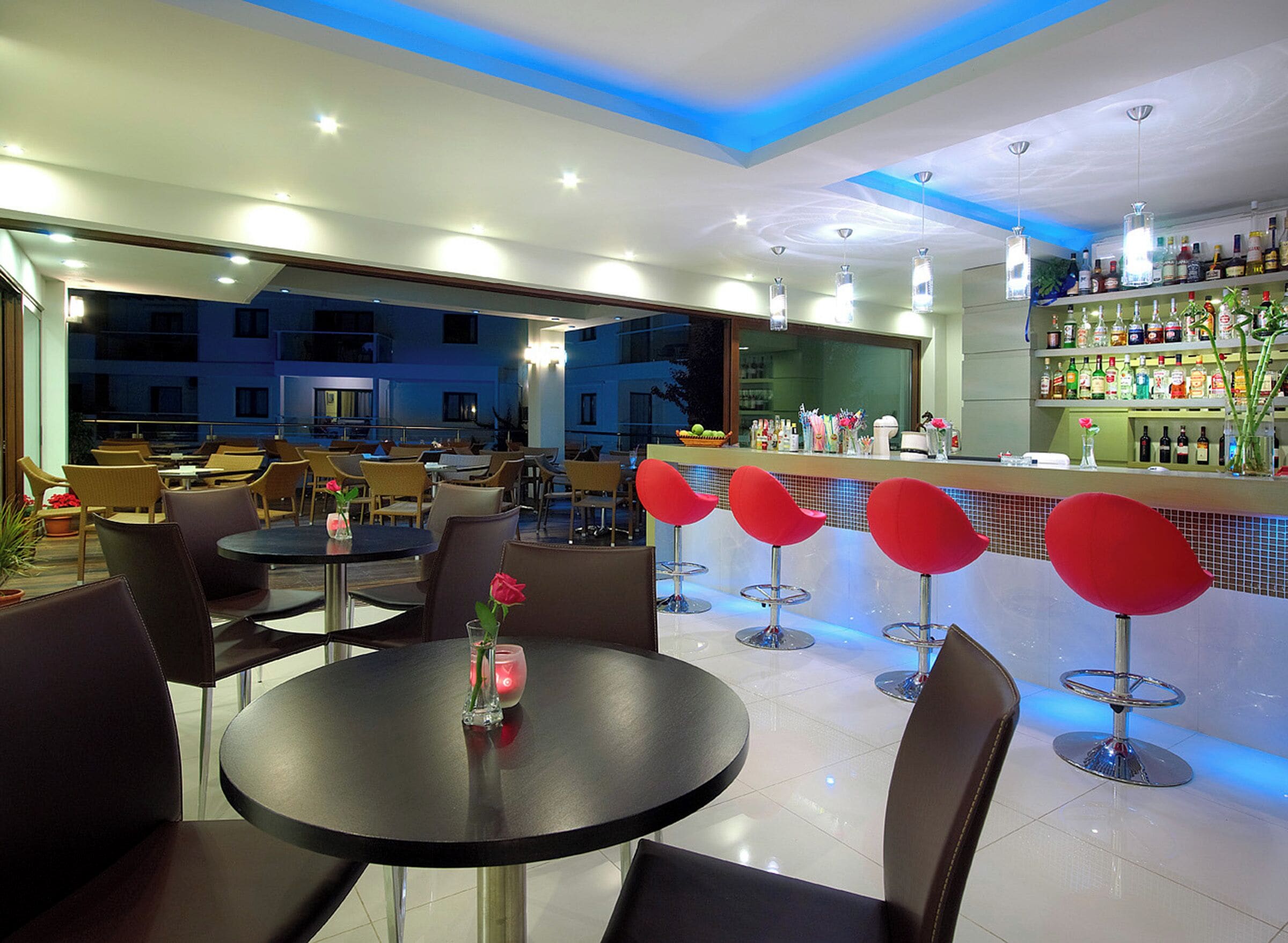 A modern bar area with round tables and red bar stools, a well-lit ceiling, and a view of the cityscape.