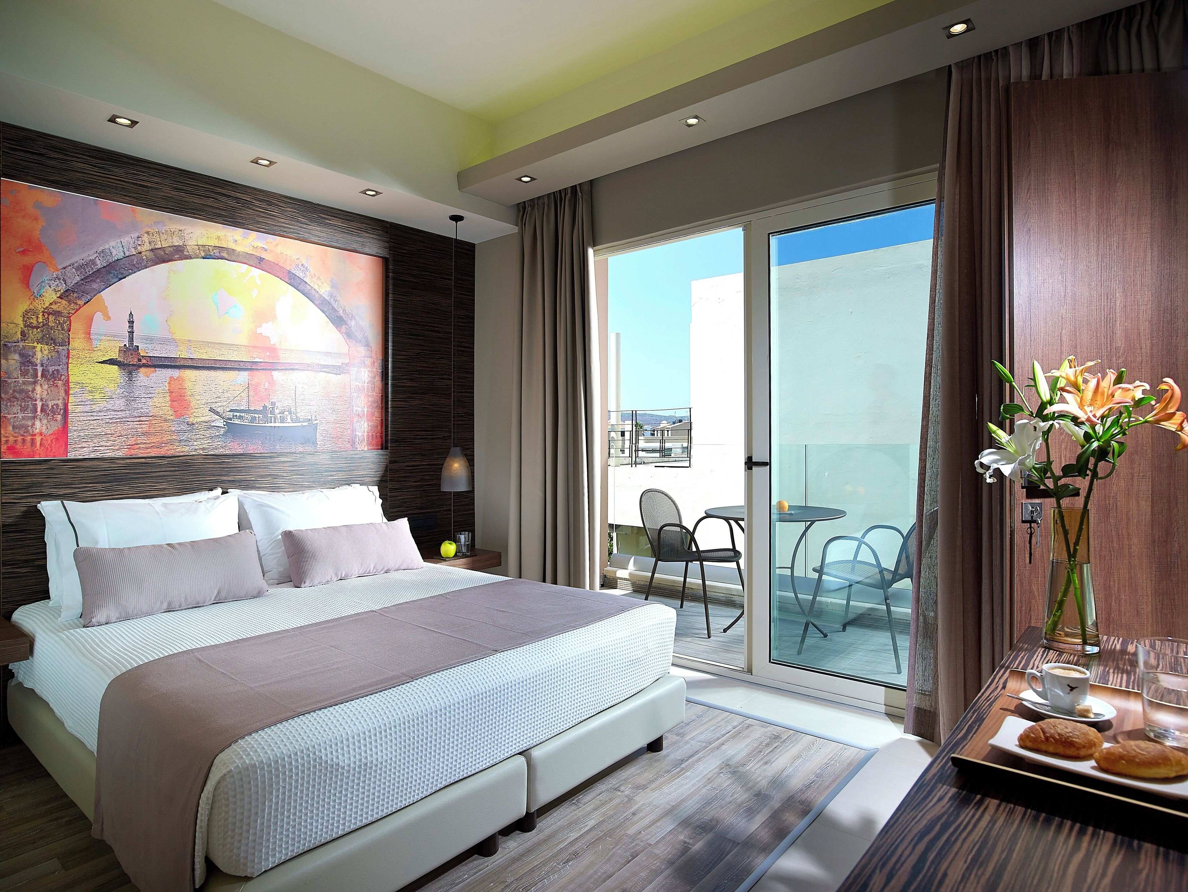 A modern hotel room with a large bed, a painting of a bridge, a balcony with a table and chairs, and a vase with flowers.
