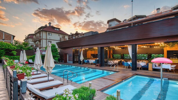 Un área de piscina en la azotea con sillones y sombrillas, una zona de bar con mesas y sillas, y vistas de edificios circundantes.
