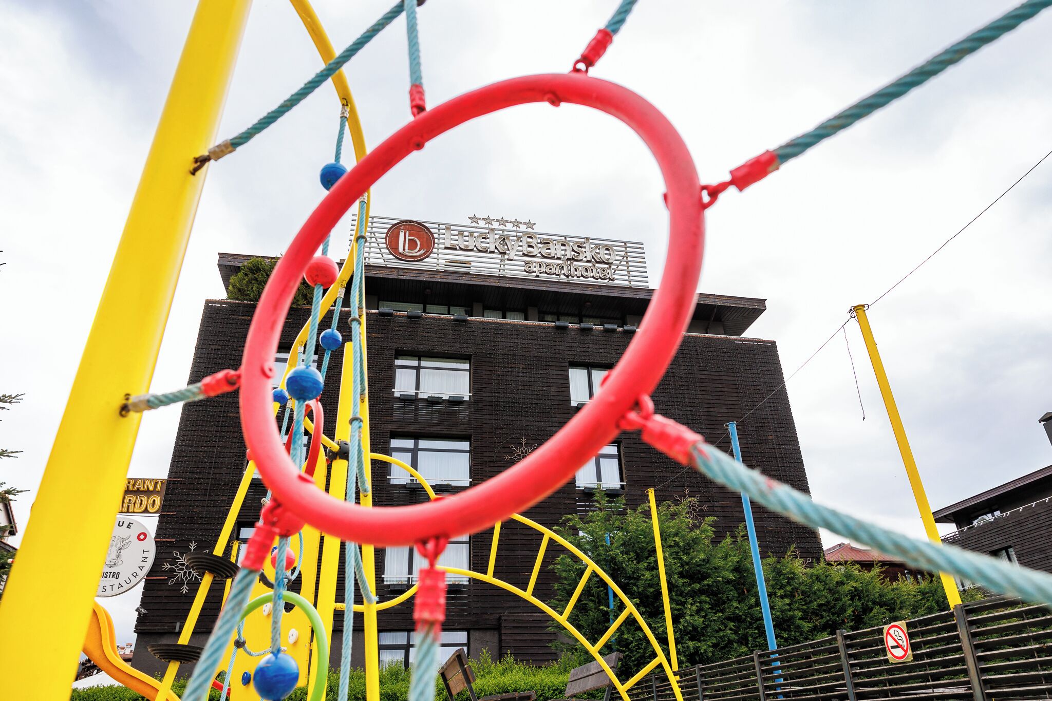 A playground structure with a red circular frame, yellow and blue ropes, and a building with a sign that reads "Lucky Barske apoteket" in the background.