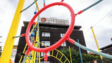 A playground structure with a red circular frame, yellow and blue ropes, and a building with a sign that reads "Lucky Barske apoteket" in the background.