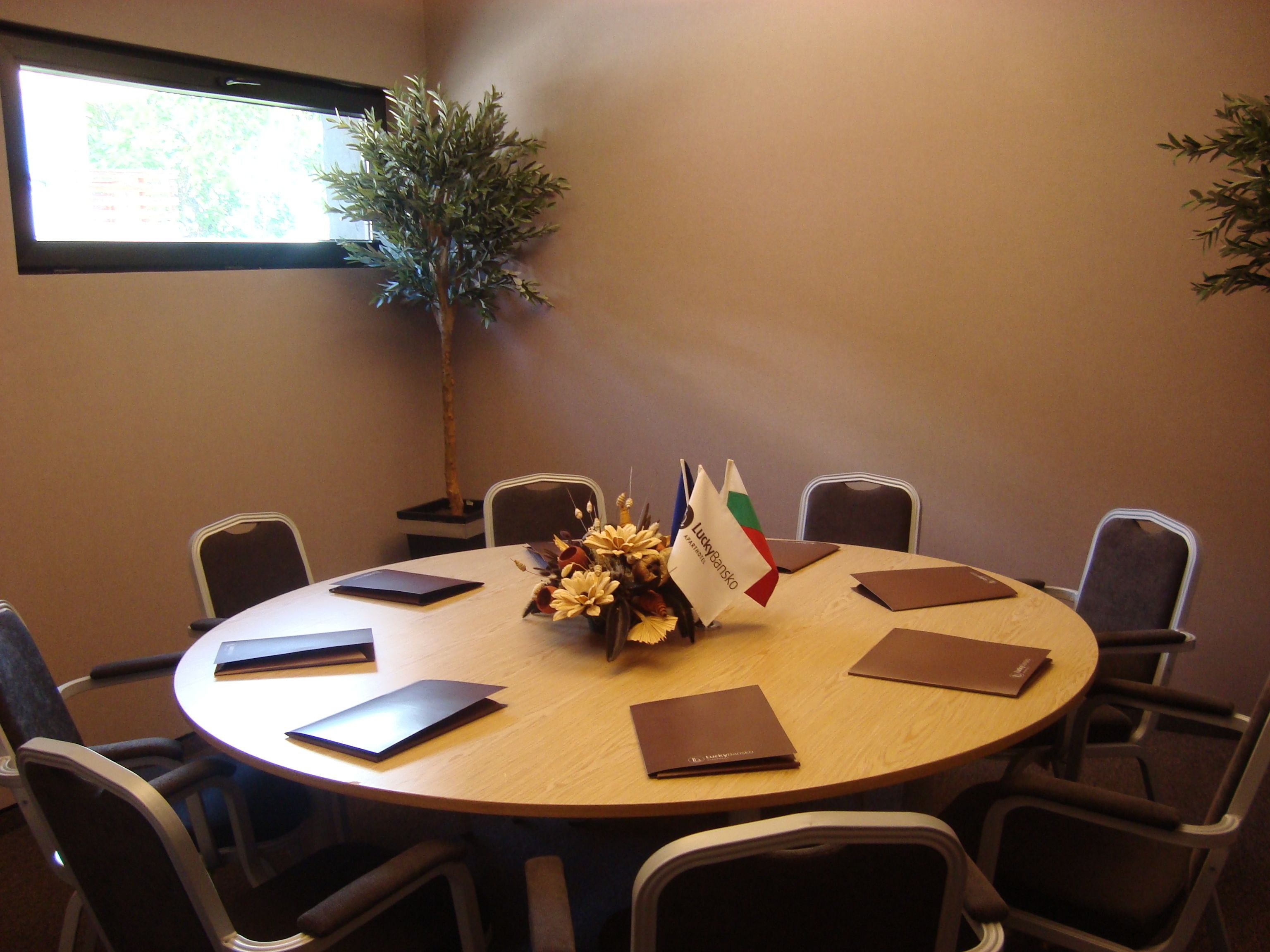 A conference room with a round table, chairs, a potted plant, and a window with blinds.