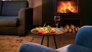 Una sala de estar acogedora con chimenea, un sofá, una mesa de comedor con fruta y una copa de vino, y una vista panorámica de árboles y montañas a través de una ventana.