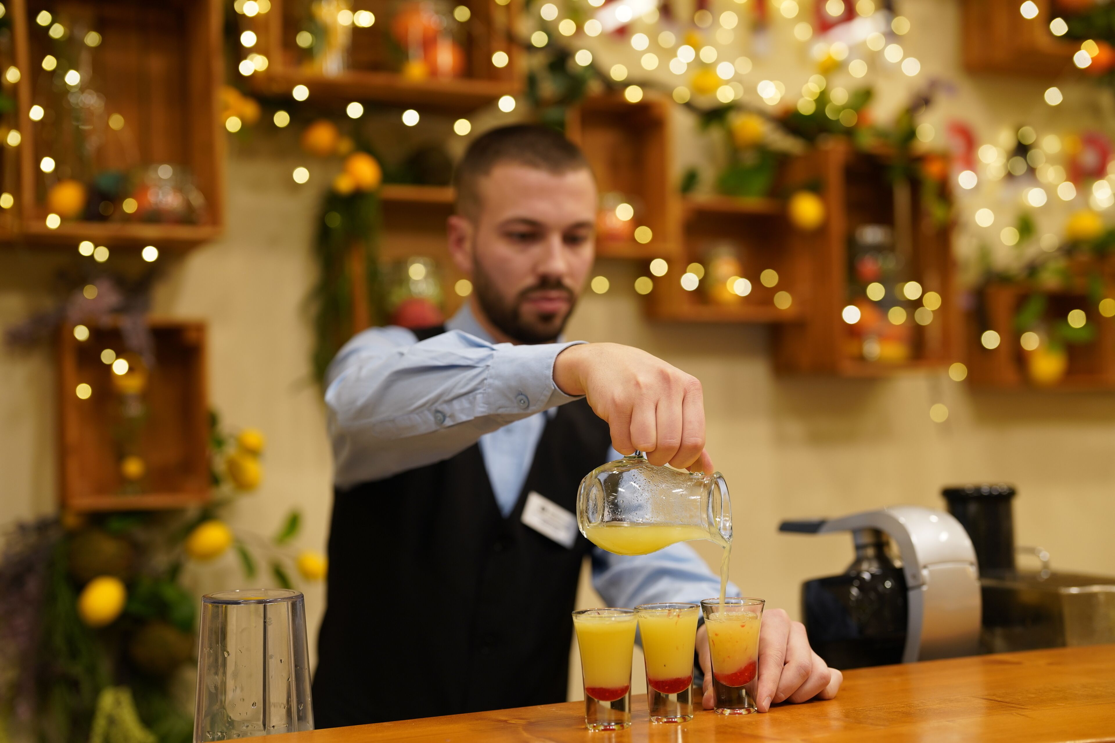 Una persona sirviendo una bebida en un vaso en un bar, con un mostrador de madera y estanterías al fondo.