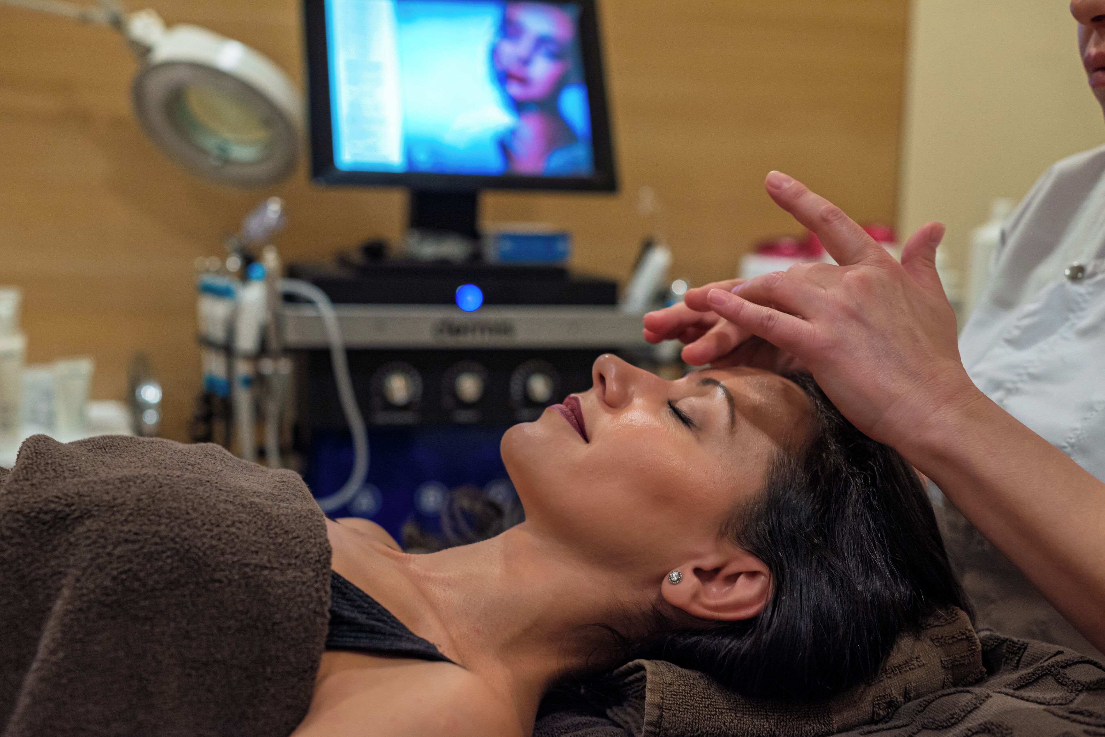 A person lying down with a medical device attached to their head, a monitor in the background, and a person standing beside them.