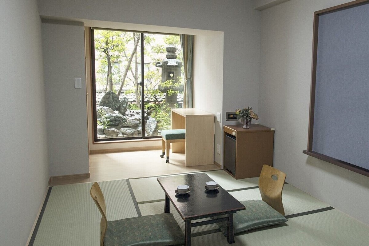 Une pièce de style japonais traditionnel, avec un sol recouvert de tatamis, une petite table basse avec des coussins, un bureau et une vue sur un jardin, accessible par une porte coulissante.