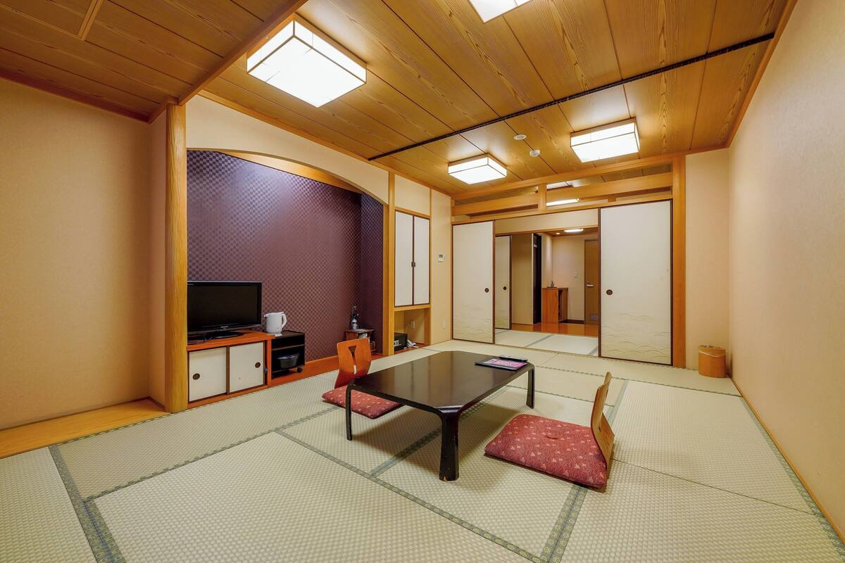 Ein traditioneller japanischer Raum mit Tatami-Boden, einem niedrigen Tisch, einem Fernseher und Schiebetüren.