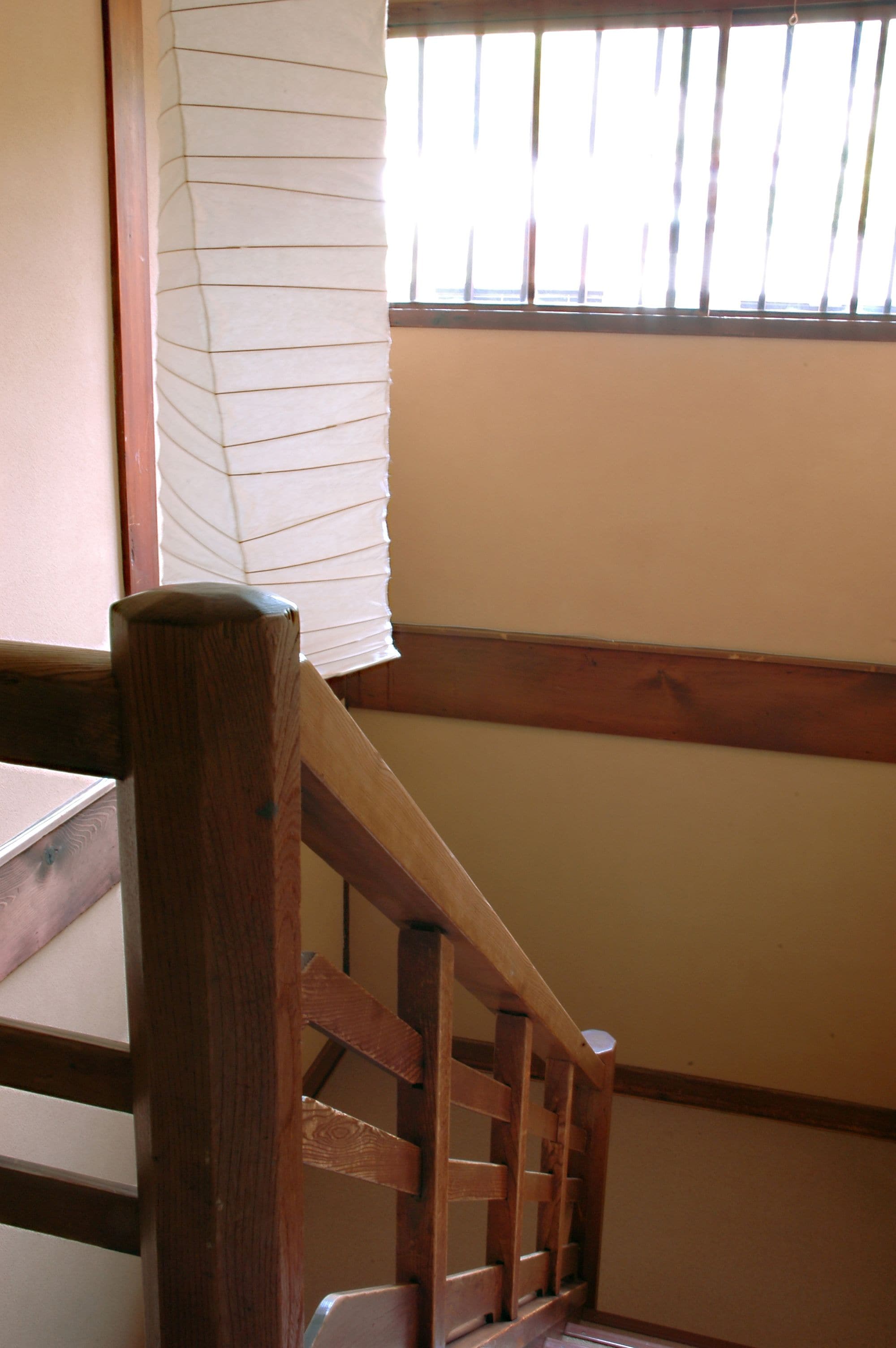A wooden staircase with a handrail, a window with blinds, and a wall with a wooden trim.