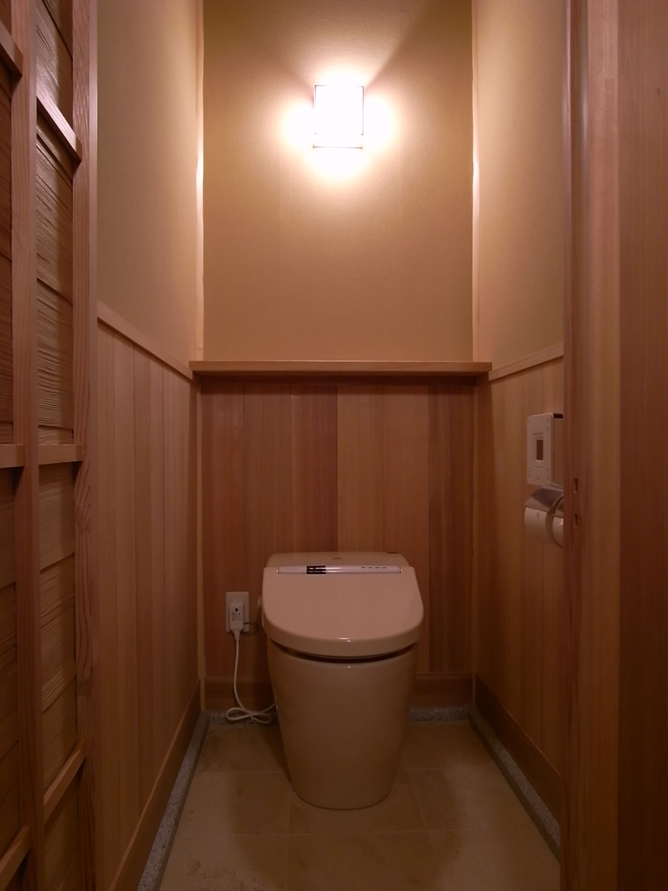 A small, wooden-paneled bathroom with a toilet, a wall-mounted light, and a visible electrical outlet.