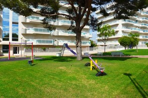A playground with a slide, swings, and a tree in front of a multi-story building with balconies.