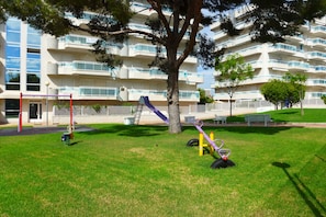 A playground with a slide, swings, and a tree in front of a multi-story building with balconies. - Apartamentos Larimar - Families Only (Salou)