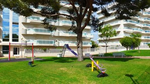Un parque infantil con tobogán, columpios y un árbol frente a un edificio de varios pisos con balcones.