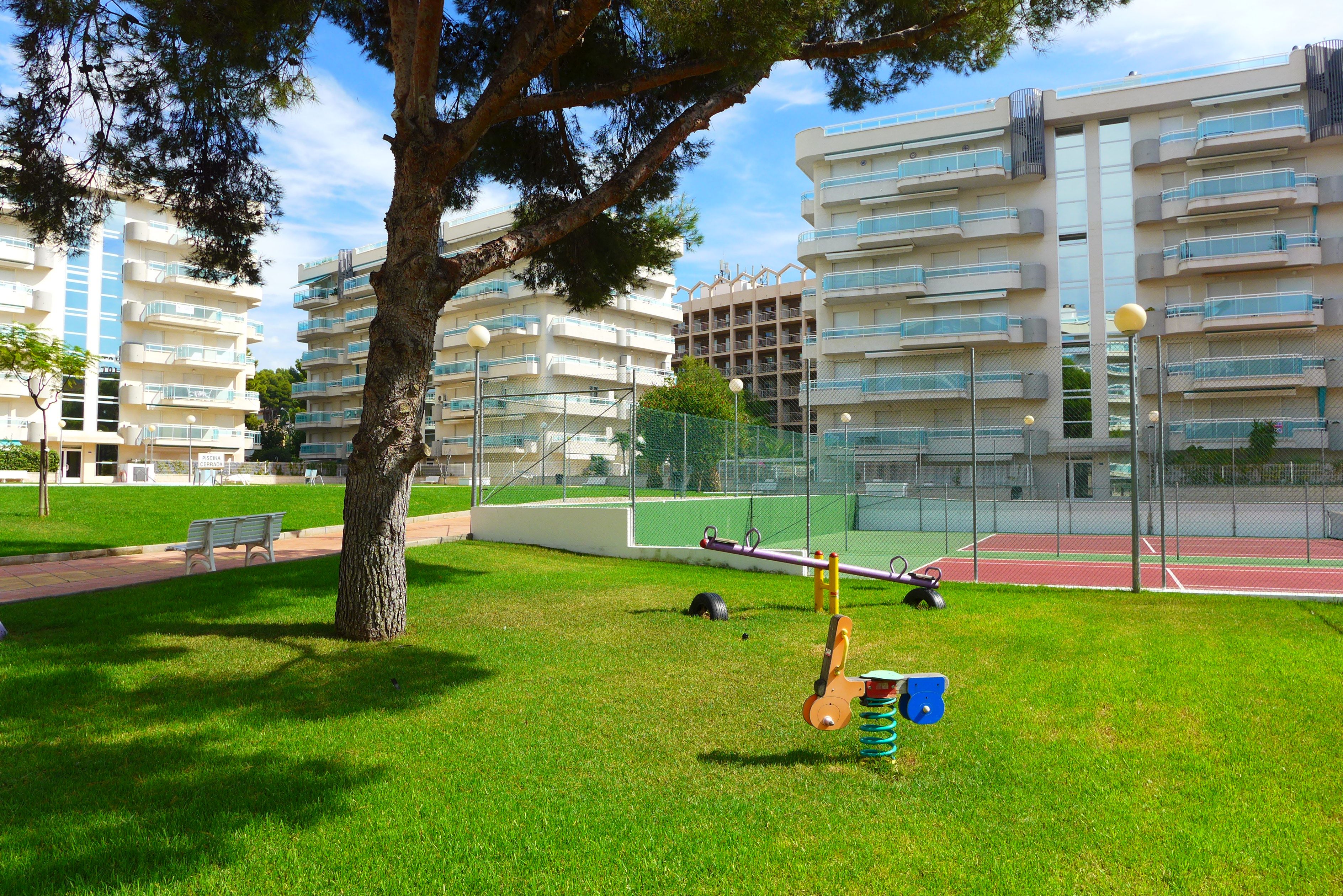 Un parc avec une aire de jeux, un arbre et un court de tennis, le tout entouré d’immeubles d’habitation.
