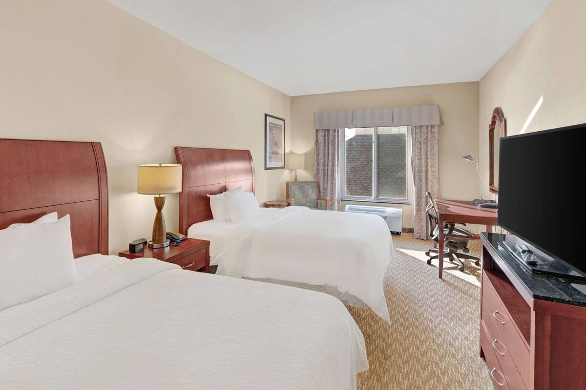 A hotel room with two beds, a desk, a television, and a window with curtains.