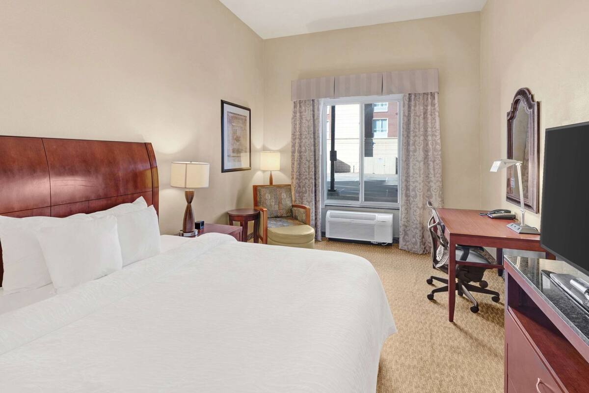 A hotel room with a large bed, a desk with a computer, a chair, a TV, and a window with curtains.