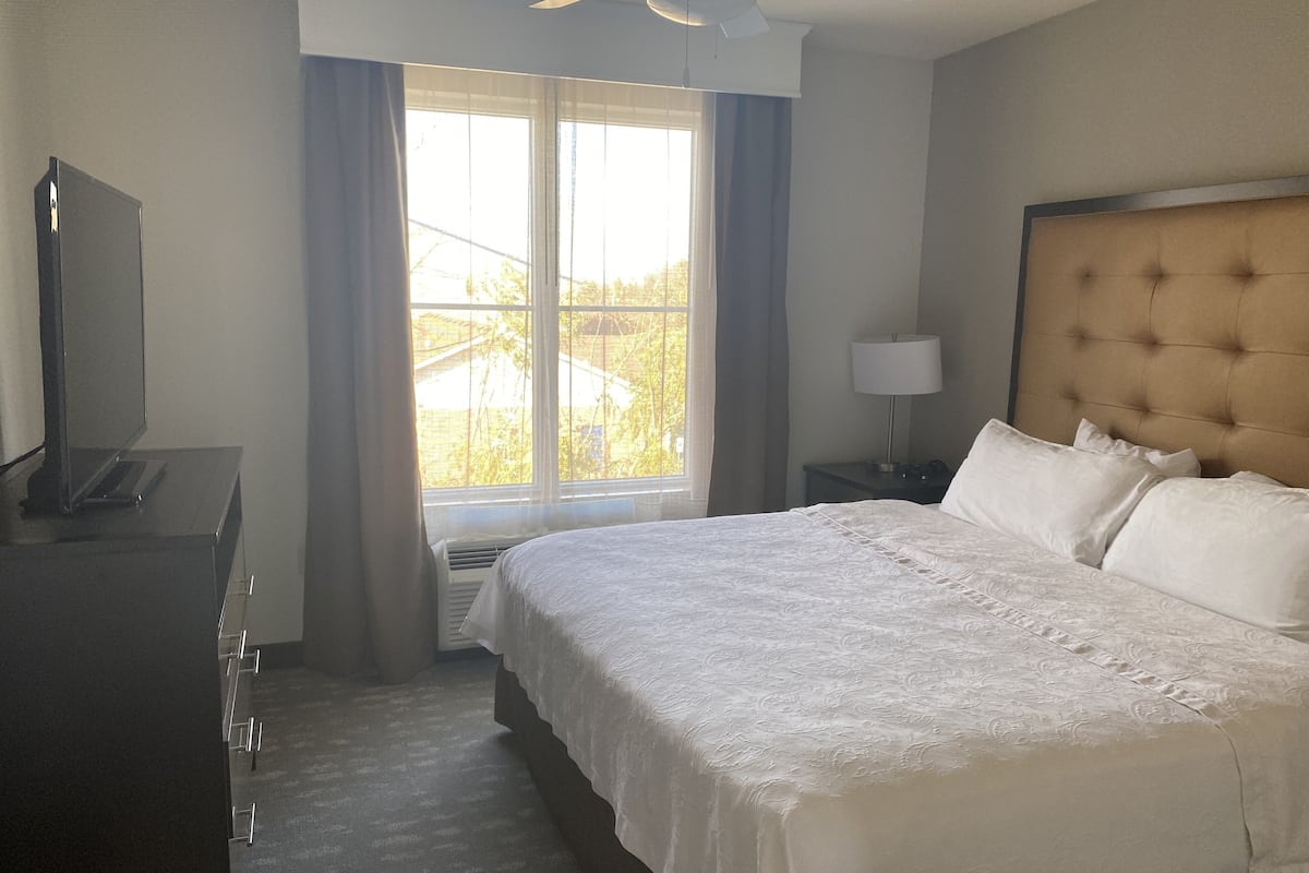 A hotel room with a large bed, a TV on a dresser, a window with curtains, and a ceiling fan.
