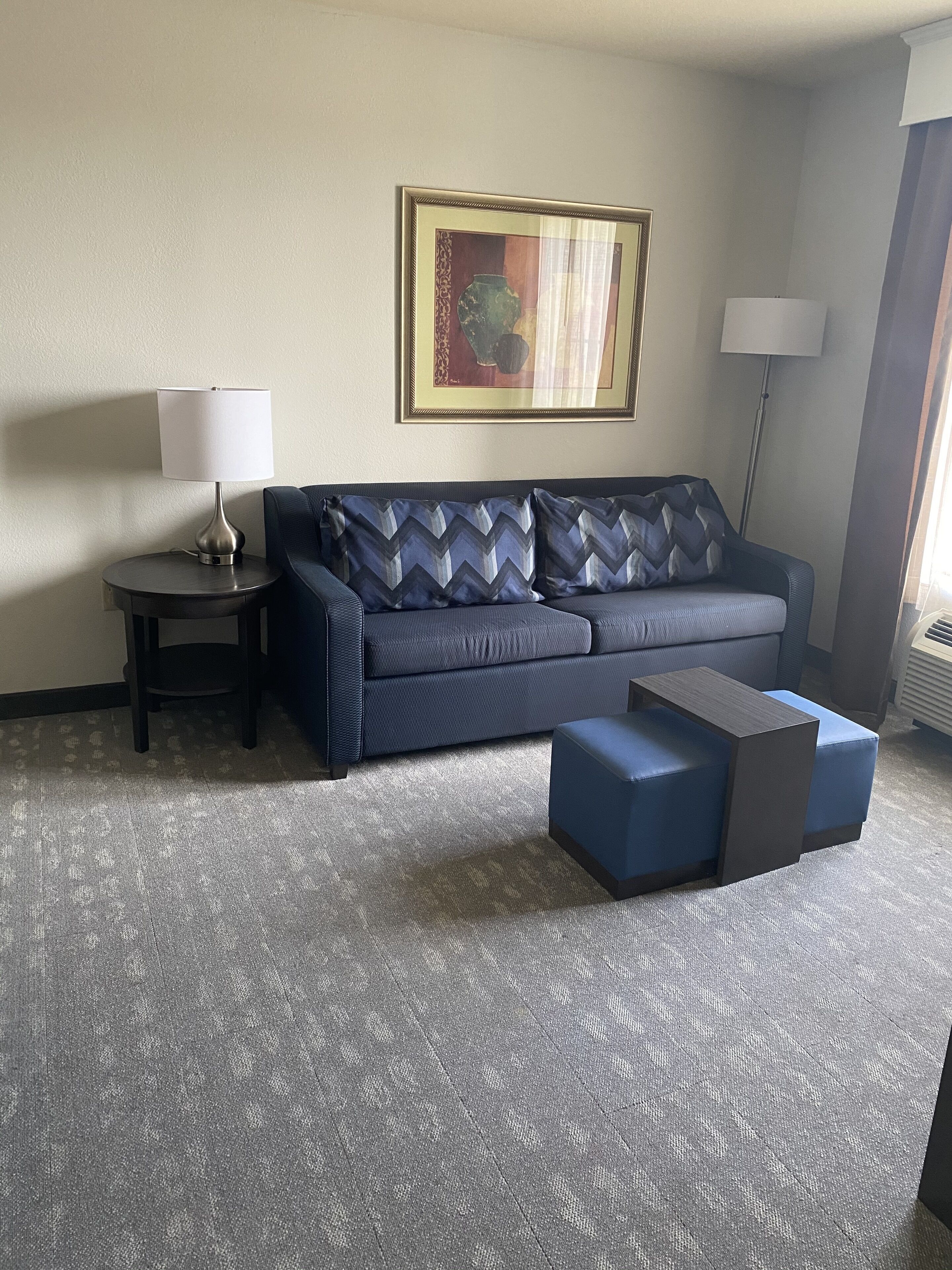 Quarto de hotel moderno com um sofá cinzento, uma pequena mesa de apoio com uma luminária, uma obra de arte emoldurada na parede e um abajur.