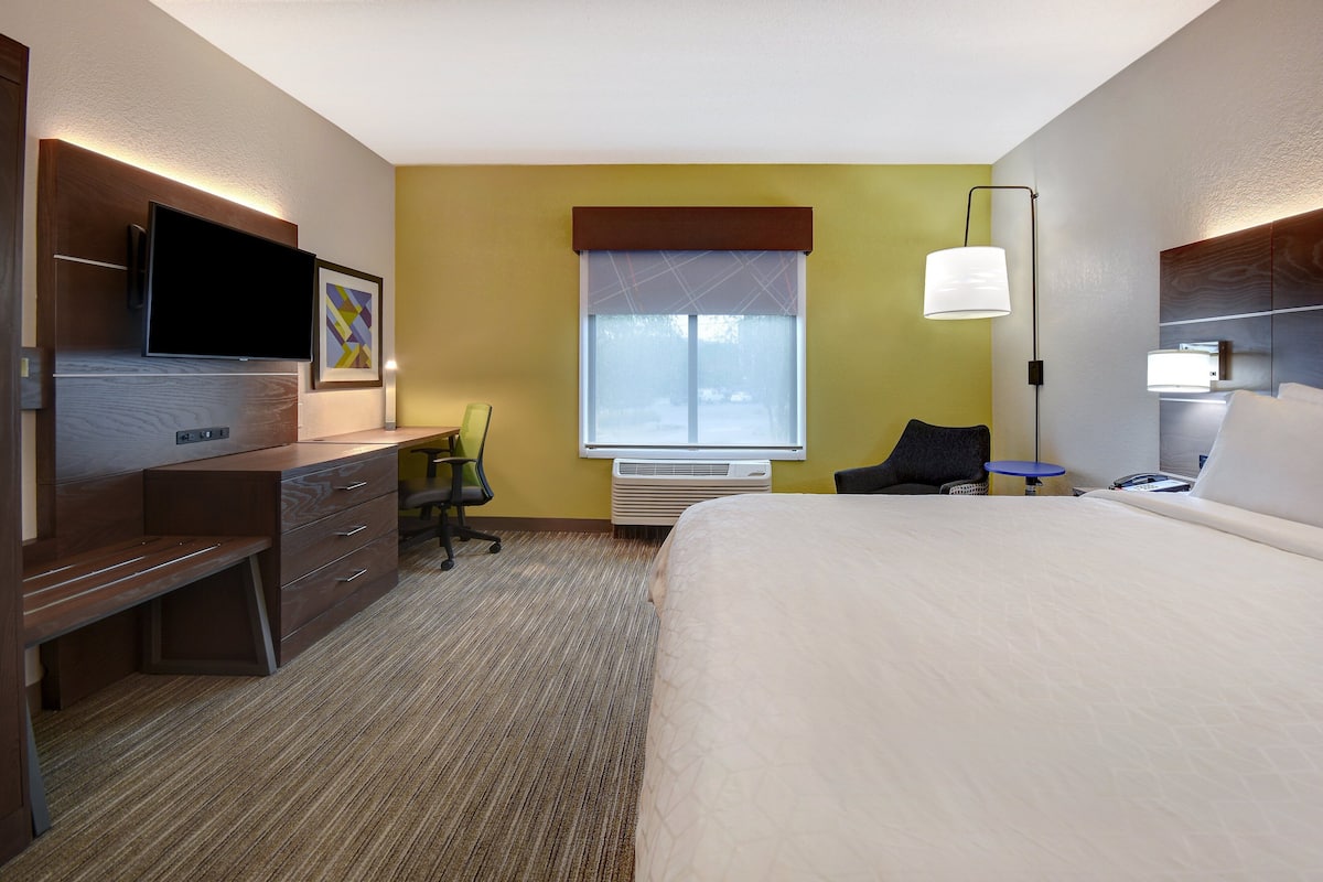 A hotel room with a bed, a desk with a flat-screen TV, a chair, a lamp, and an air conditioning unit.