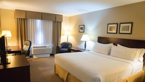 A hotel room with a large bed, a desk with a computer, a chair, a lamp, and a window with curtains.