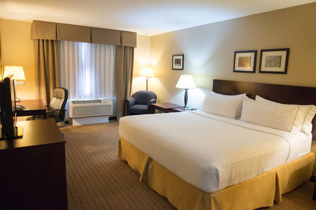 A hotel room with a large bed, a desk with a computer, a chair, a lamp, and a window with curtains.