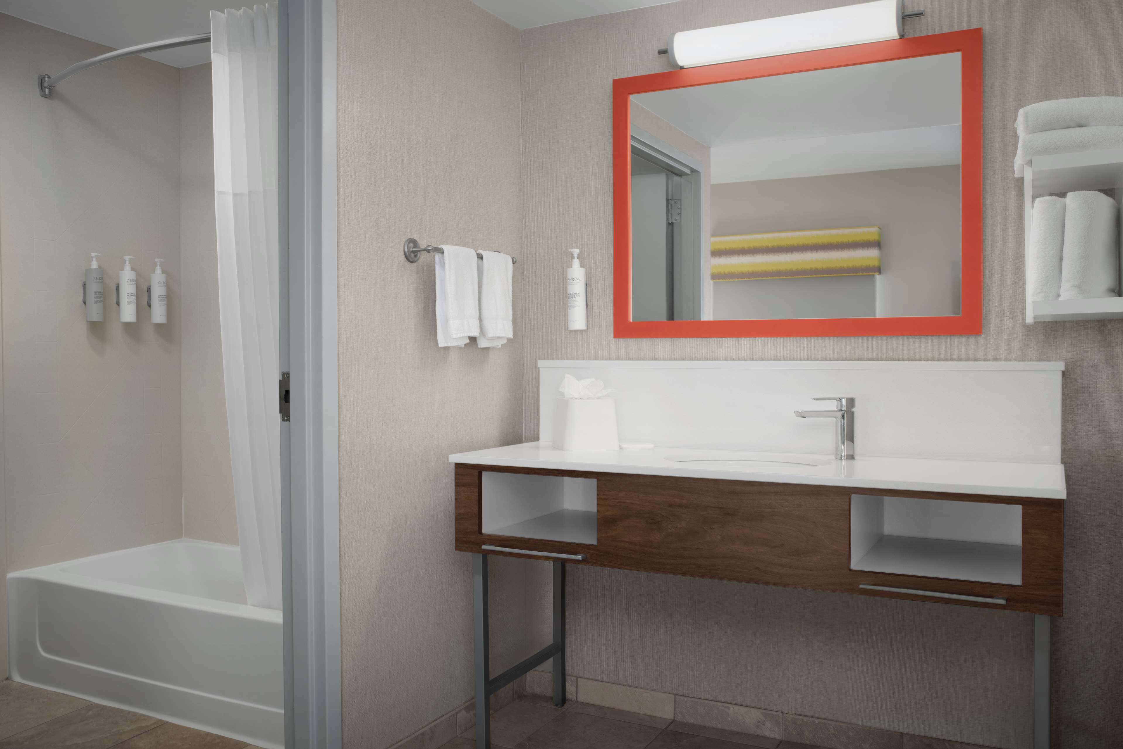Un baño moderno con ducha, un lavamanos blanco con tocador de madera, un espejo con marco rojo y un perchero para toallas.