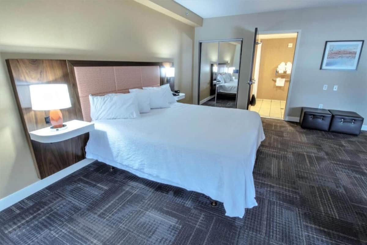 A hotel room with a large bed, a bedside table with a lamp, a wardrobe, and a bathroom visible through an open door.