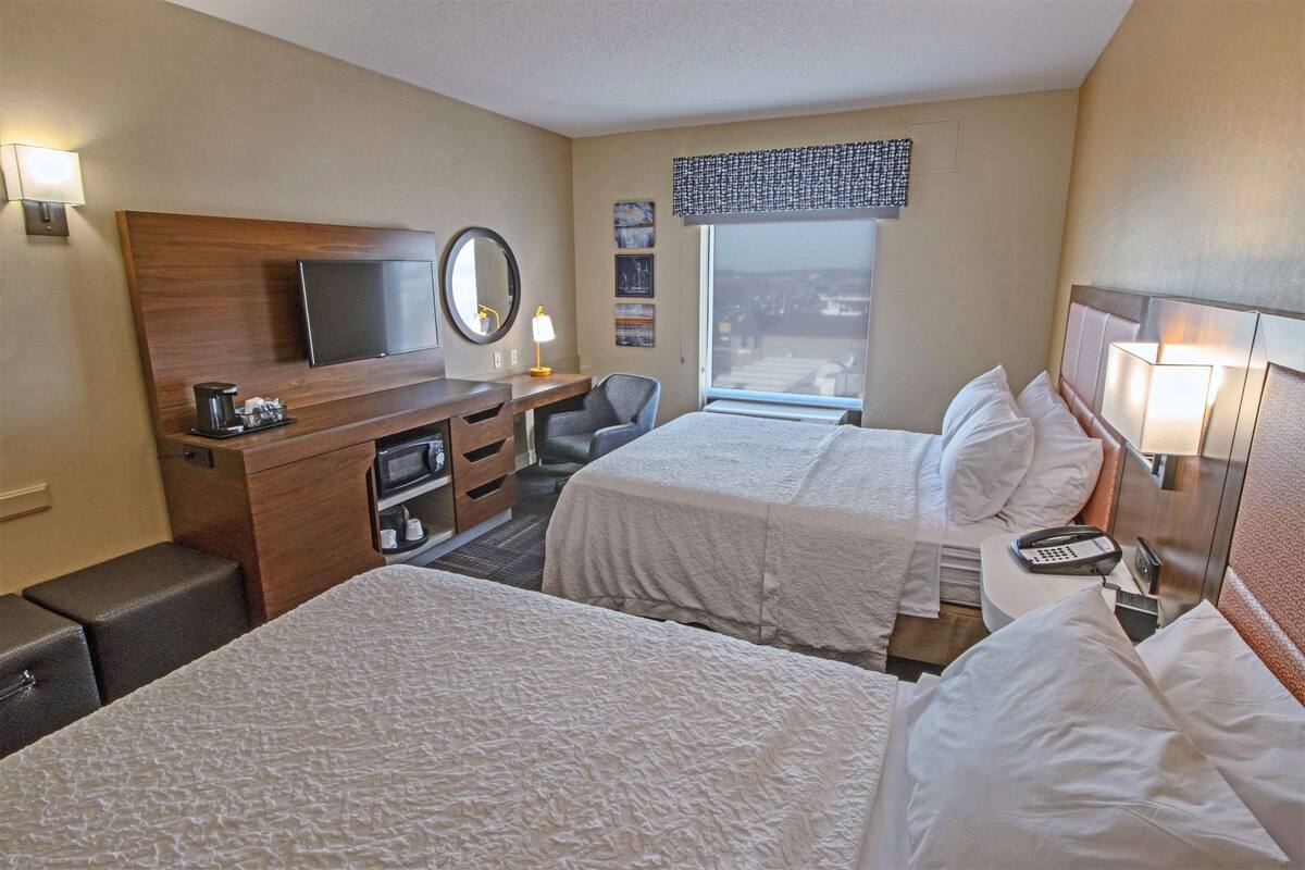 A hotel room with two beds, a desk, a chair, a TV, a window with a view, and a bedside table with a lamp.