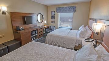 A hotel room with two beds, a desk, a chair, a TV, a window with a view, and a bedside table with a lamp.