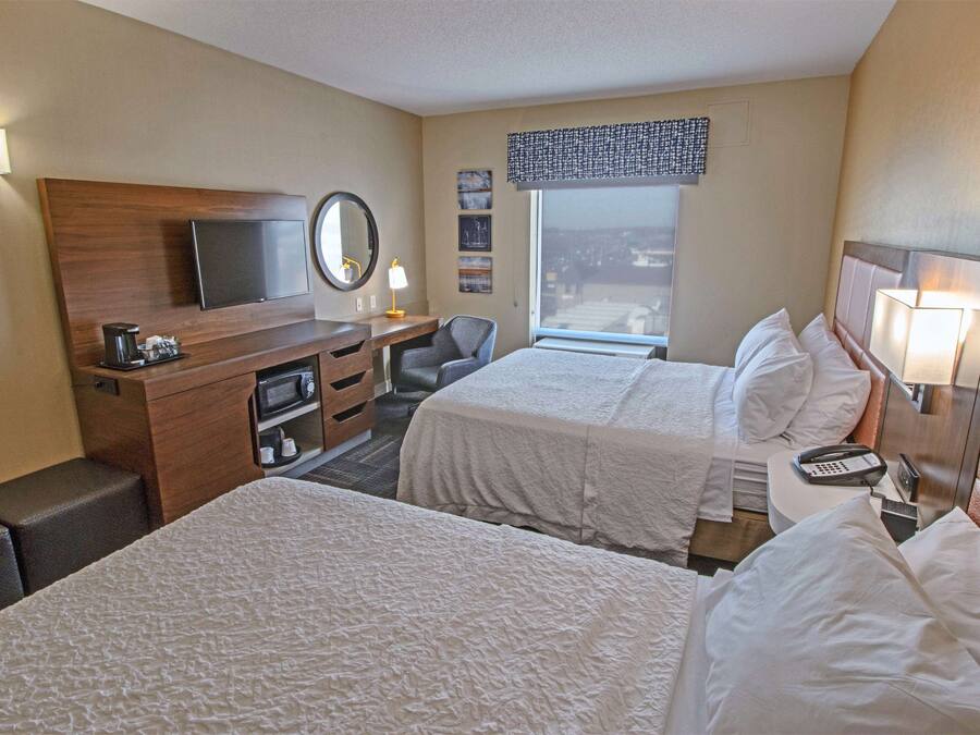 A hotel room with two beds, a desk, a chair, a TV, a window with a view, and a bedside table with a lamp.