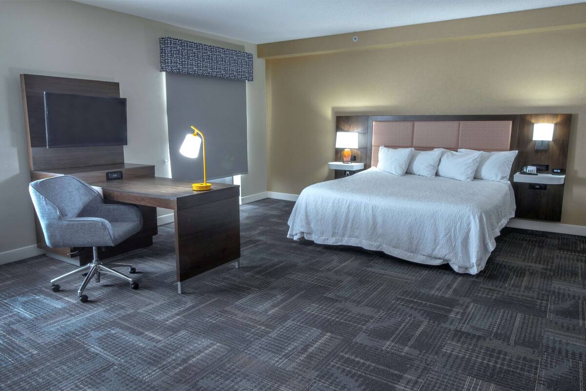 A hotel room with a bed, desk, chair, TV, lamps, and a patterned carpet.