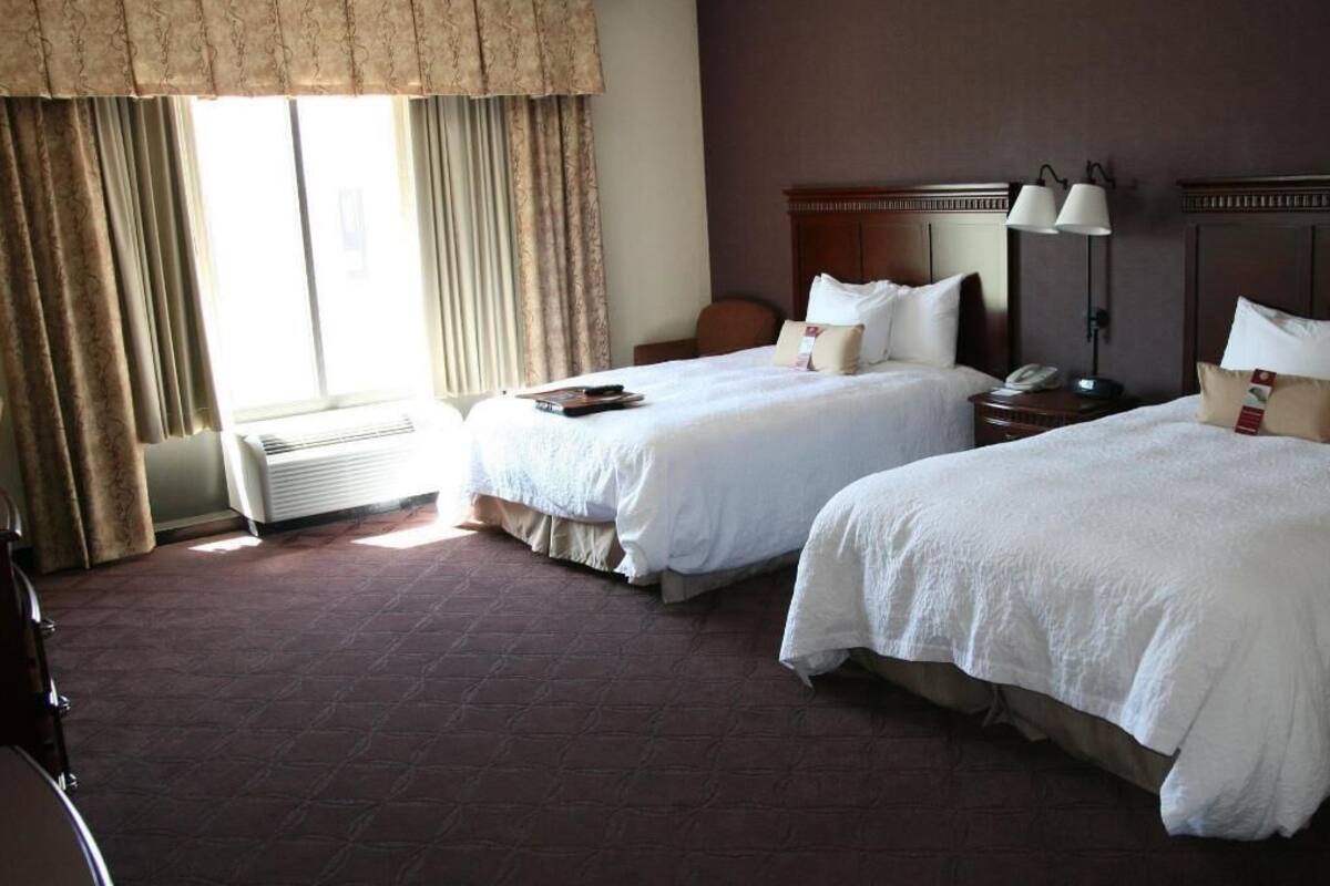 A hotel room with two beds, a desk, a chair, a lamp, and a window with curtains.