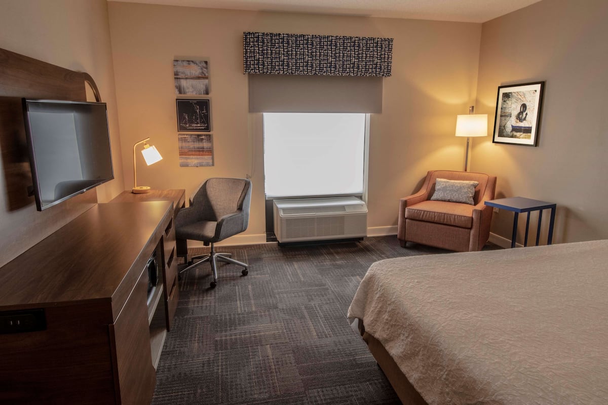 A hotel room with a bed, a desk with a flat-screen TV, a chair, a window with a view, a wall-mounted lamp, and a framed picture.