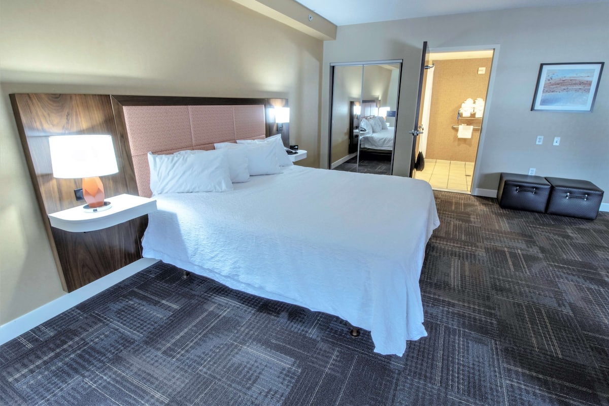 A hotel room with a large bed, a nightstand with a lamp, a wardrobe, and a bathroom visible through an open door.