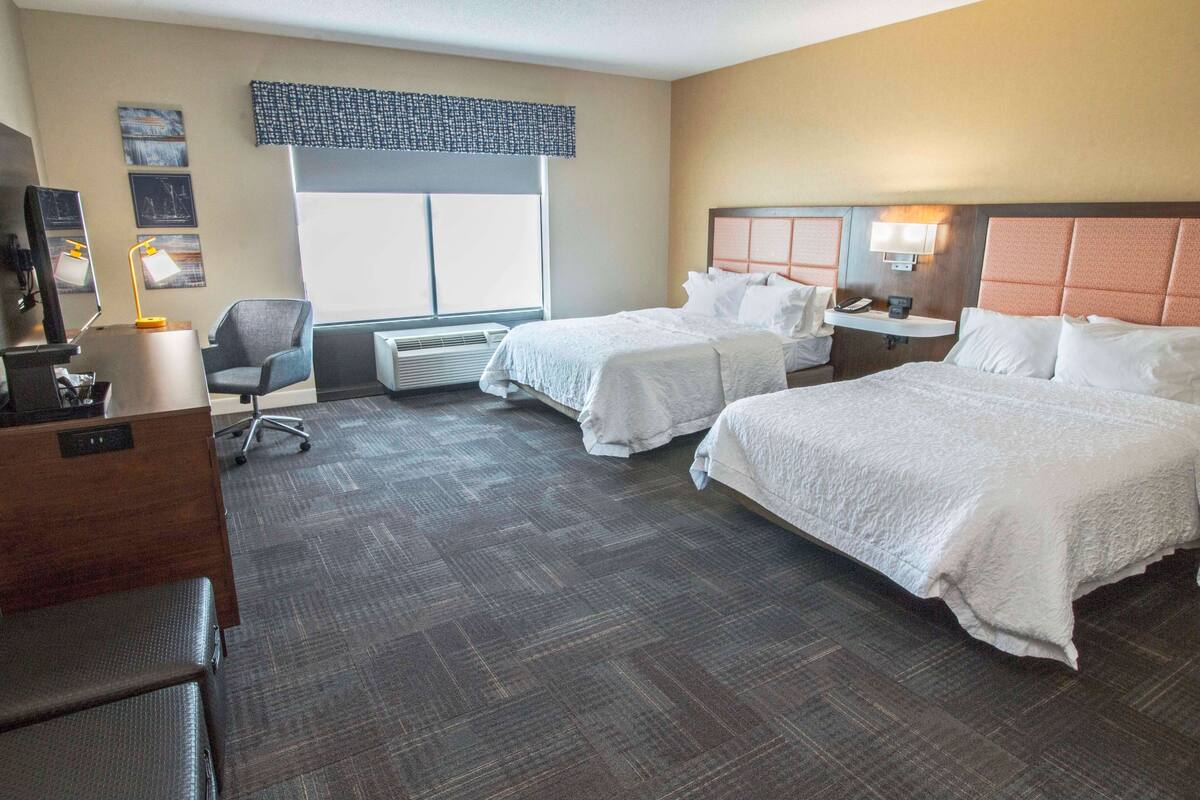 A hotel room with two beds, a desk, a chair, a desk lamp, a window with blinds, and artwork on the walls.
