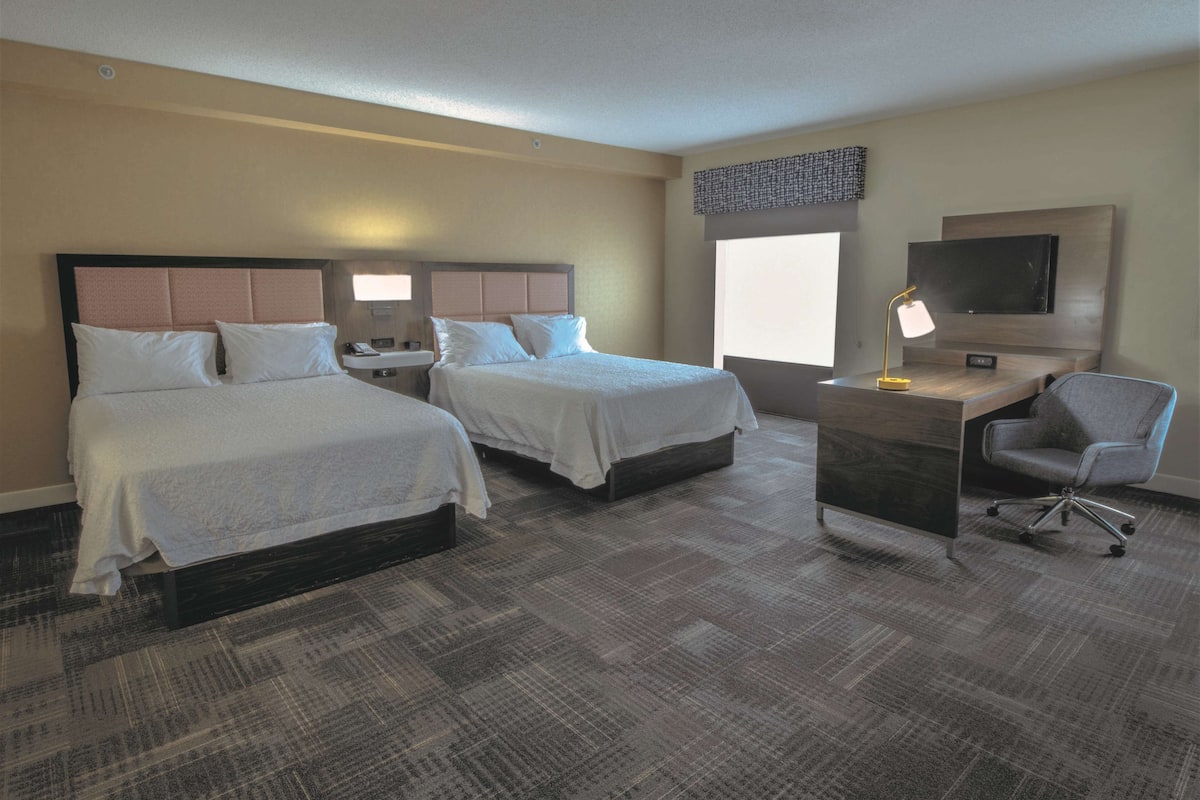 A hotel room with two beds, a desk, a chair, a TV, and a window.