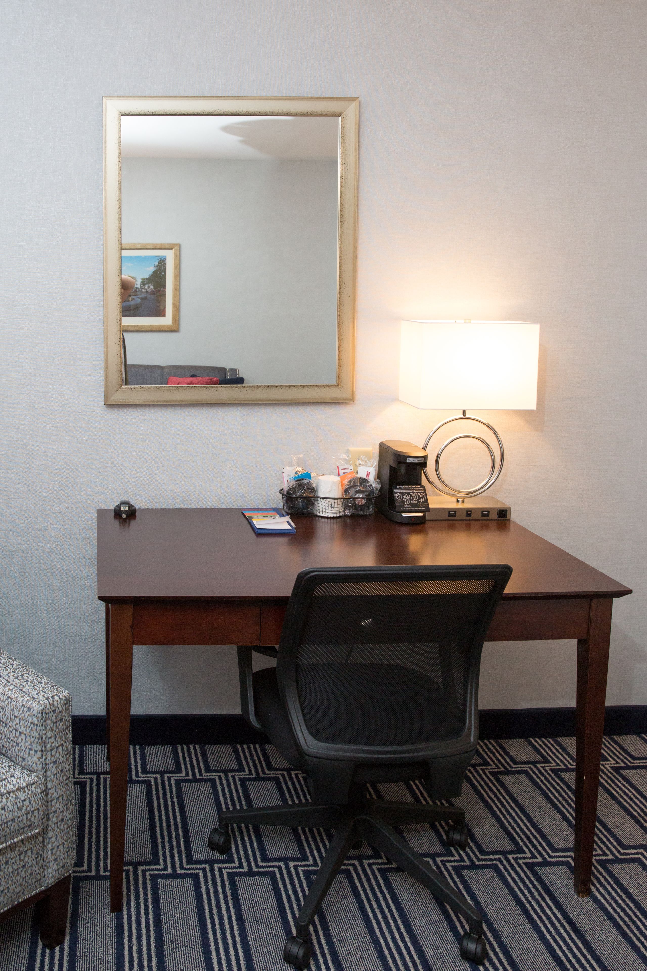 Ein Hotelzimmer mit einem Holztisch, einem schwarzen Bürostuhl, einer Lampe, einer Kaffeemaschine und einem kleinen Korb mit Toilettenartikeln.