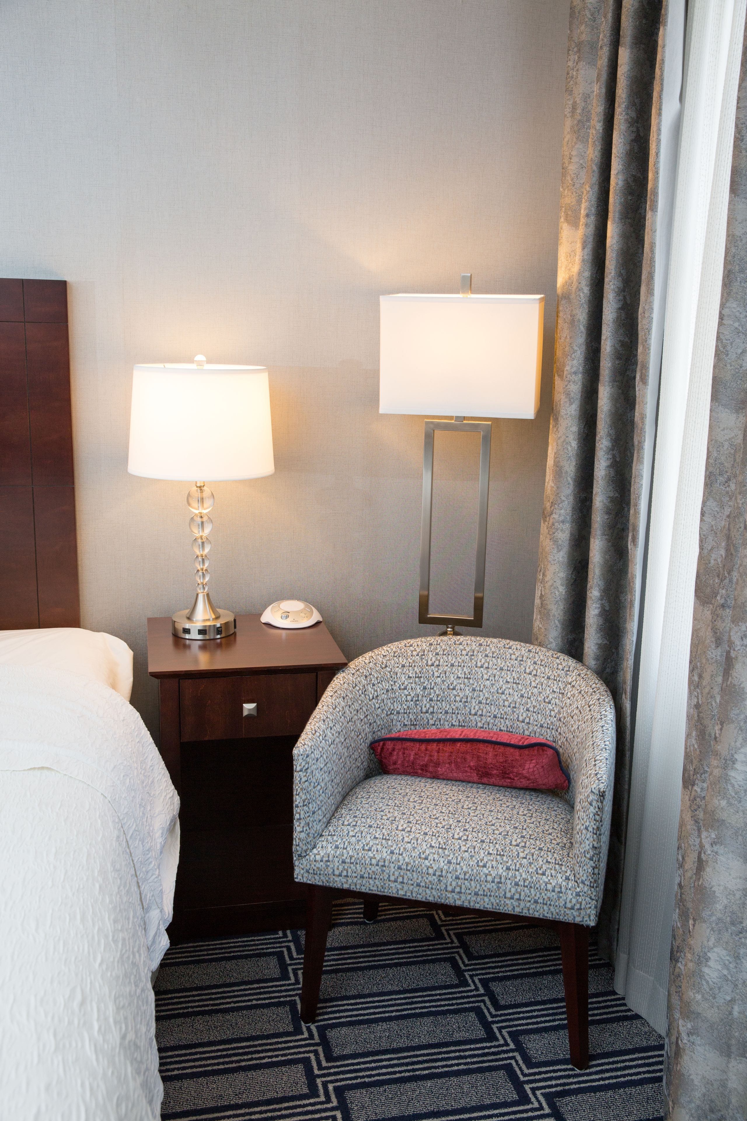 A hotel room with a bed, a desk with a lamp, a chair with a red pillow, and a patterned rug.