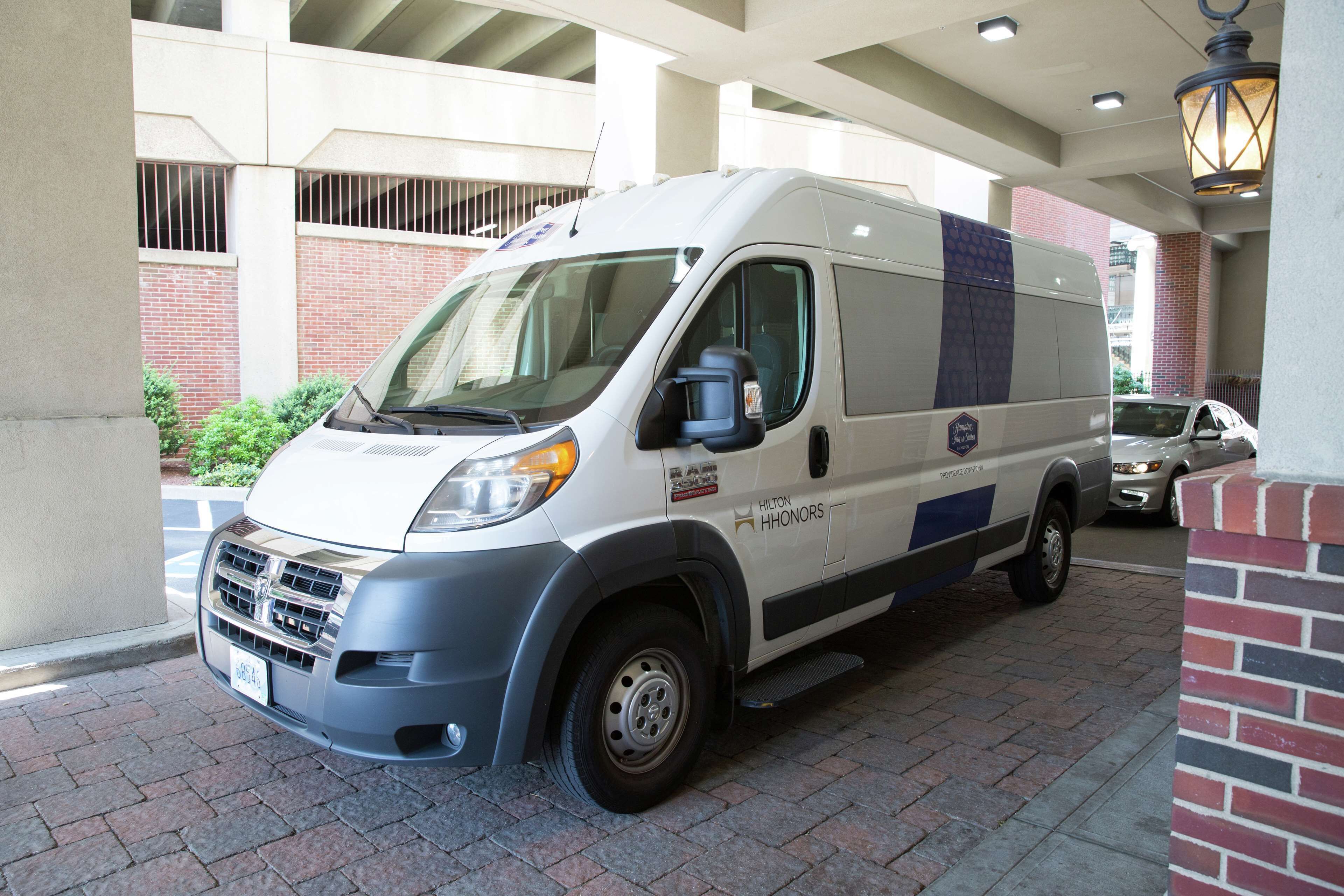 A white van parked in a covered parking area with a brick wall and a metal railing in the background.
