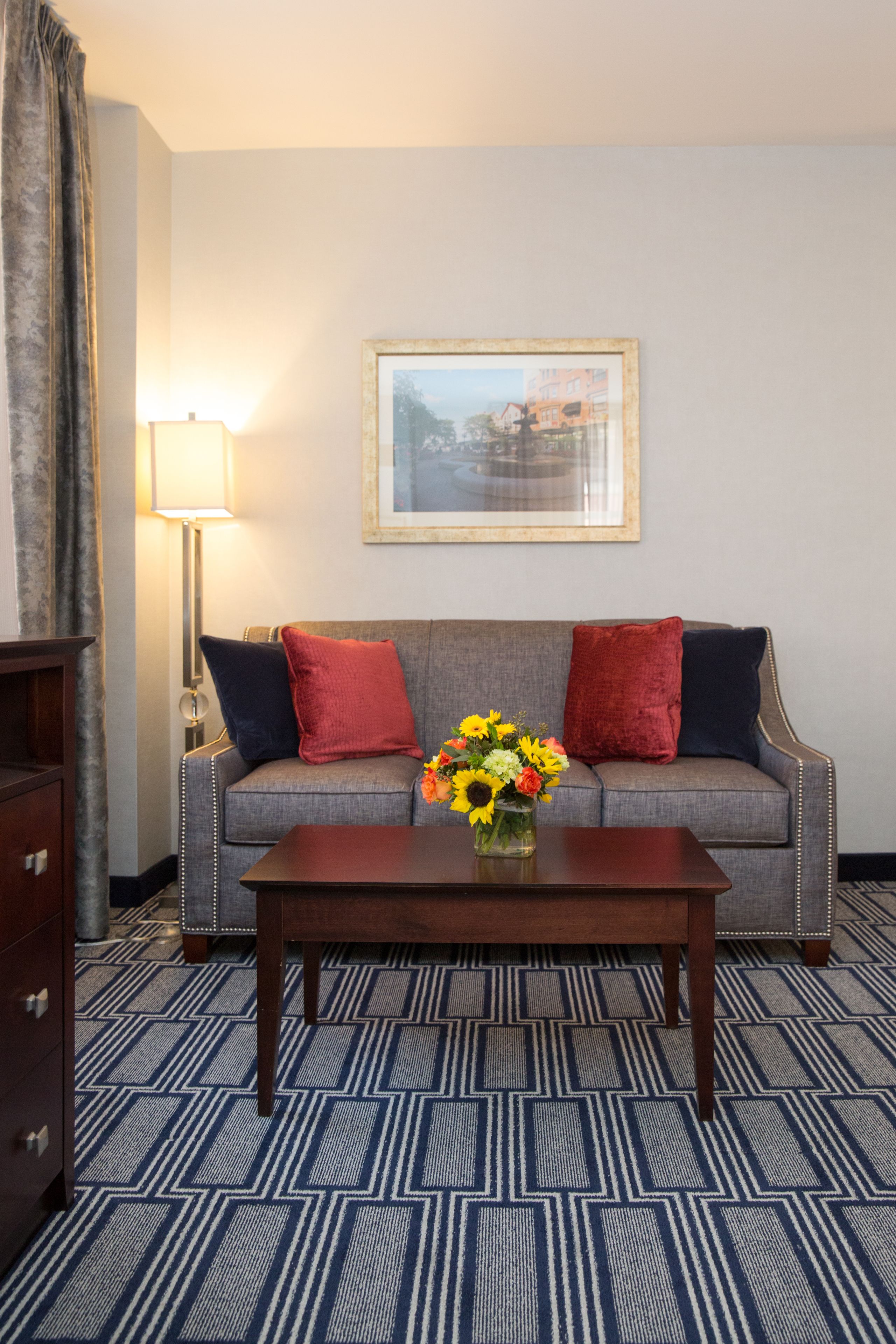 Habitación de hotel con sofá gris, dos almohadas rojas y una azul, una mesa de centro de madera, un jarrón con flores, un cuadro enmarcado en la pared y una alfombra estampada.